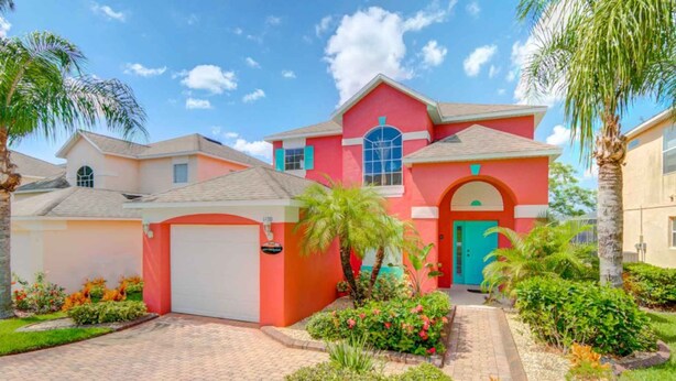 A two-story house with a red facade, a garage, and a well-maintained garden with palm trees and flowering plants. - Southern Dunes 1120 (Haines City)