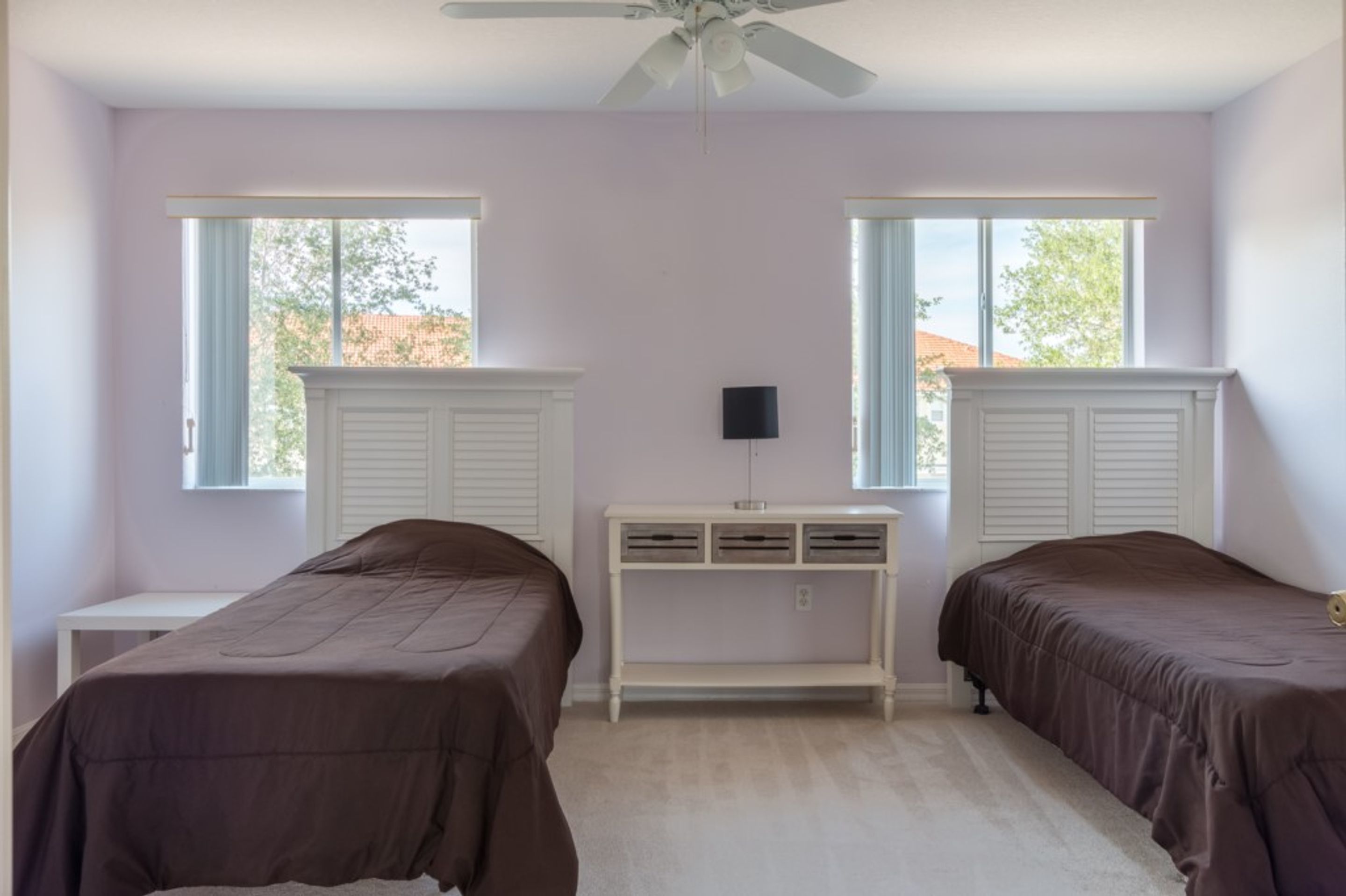 A bedroom with two beds, a white console table, a ceiling fan, and windows with shutters.