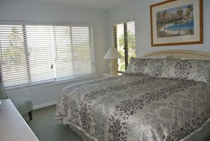 A bedroom with a large bed, a desk, and a chair. There are two windows with blinds, a lamp on a bedside table, and a framed picture on the wall.