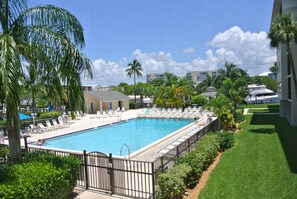 A swimming pool with lounge chairs, surrounded by greenery and palm trees, with buildings in the background.