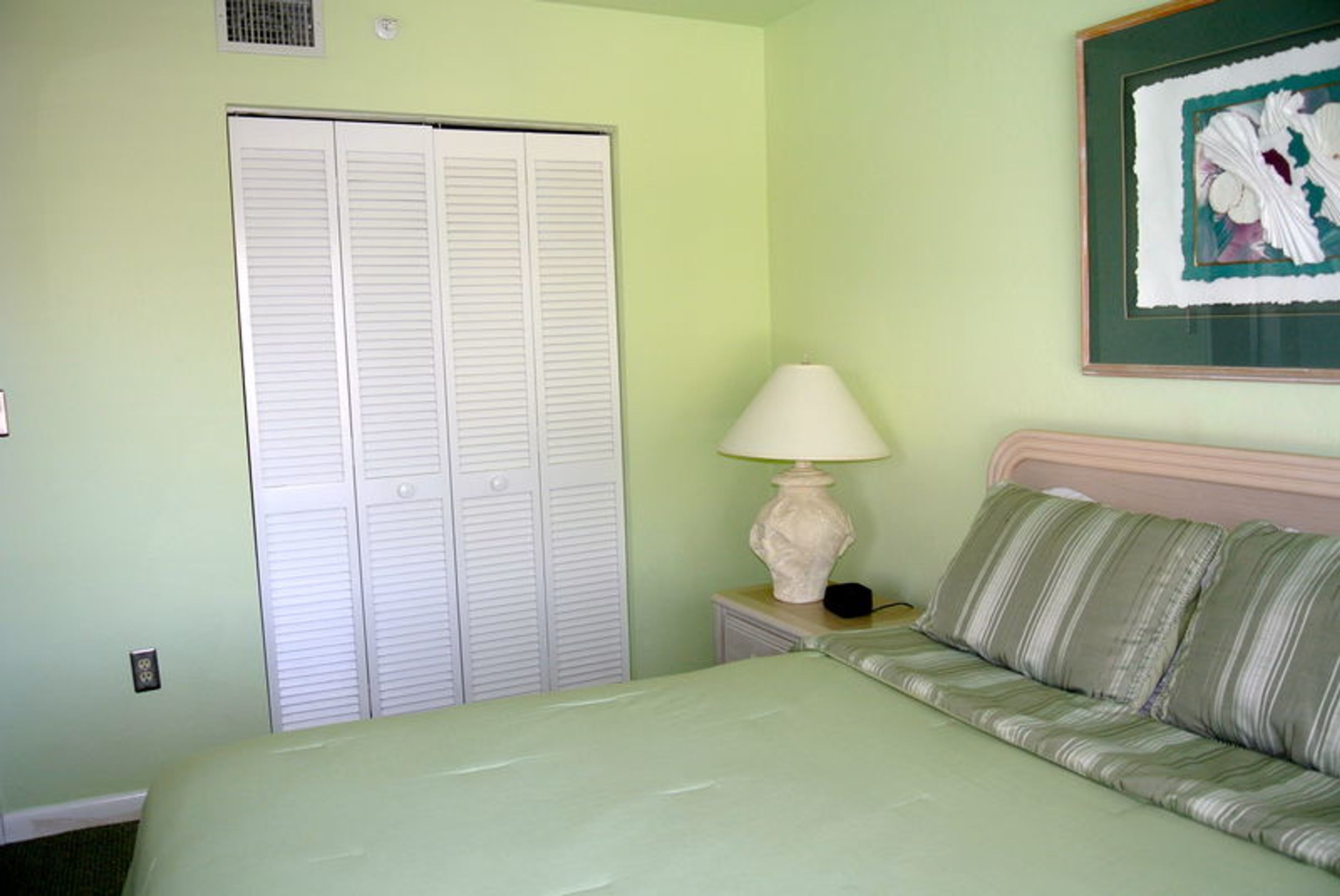 A bedroom with a bed, a nightstand with a lamp, a closet, and a framed picture on the wall.