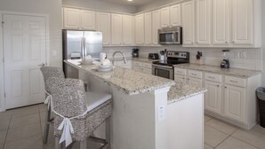 A modern kitchen with white cabinetry, a granite countertop, stainless steel appliances, and a bar area with wicker chairs. - Golden Palms Vacation Resort 2580 (Kissimmee)