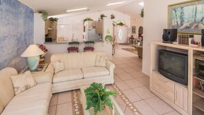 A living room with a sofa, a coffee table with a plant, a TV on a stand, and a kitchen in the background. - Lindfields 2952 (Kissimmee)