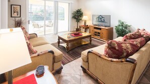 A living room with a sofa, armchairs, a coffee table, a TV, and a sliding glass door leading to an outdoor area. - Calabay Parc 215 (Davenport)