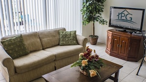 A living room with a beige sofa, a wooden coffee table with a floral arrangement, a potted plant, and a television displaying 'Florida Vacation Homes'. - Windsor Hills 2573 (Kissimmee)