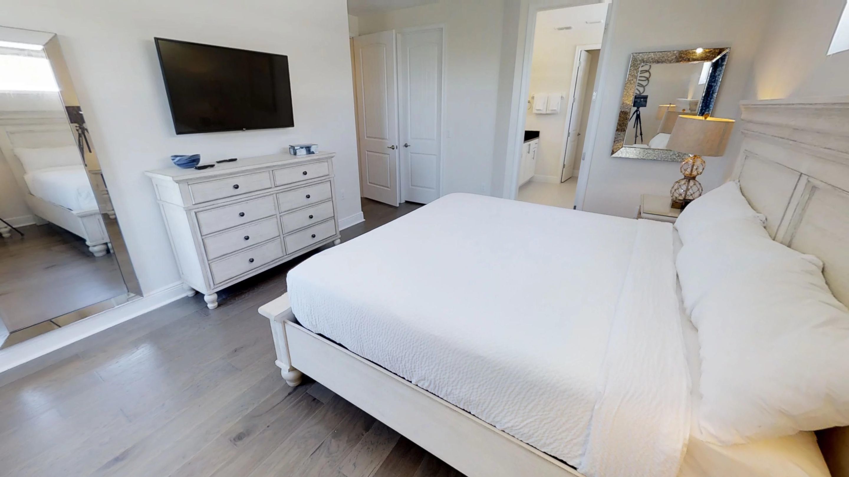 A bedroom with a large bed, a dresser, a TV, and a bathroom visible in the background.