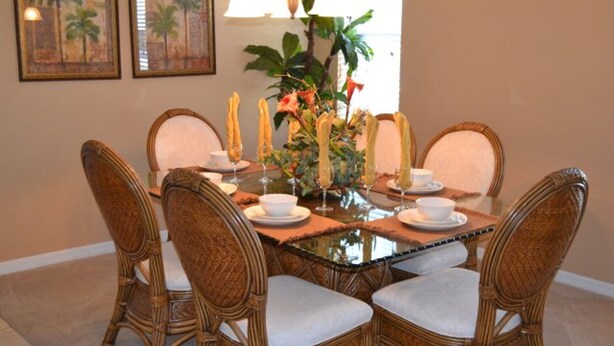 A dining table with six wicker chairs, a glass top, and a centerpiece with tall candles and flowers.