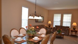A dining area with a glass-top table, wicker chairs, and a chandelier. There is a sofa and a coffee table in the background.