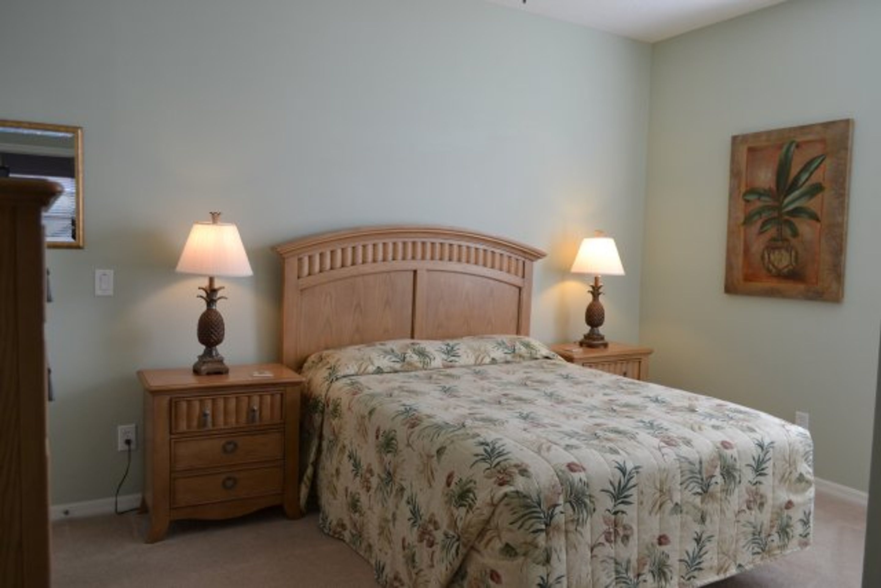 Un dormitorio con cabecera de madera, dos mesitas de noche con lámparas, una colcha con estampado floral y un cuadro colgado en la pared.