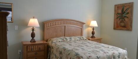 Un dormitorio con cabecera de madera, dos mesitas de noche con lámparas, una colcha con estampado floral y un cuadro colgado en la pared.
