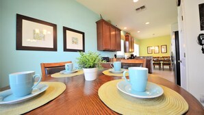 A dining area with a wooden table set for four, surrounded by chairs. There are framed pictures on the wall, a potted plant in the center, and a kitchen visible in the background. - Crystal Cove 4718 (Kissimmee)