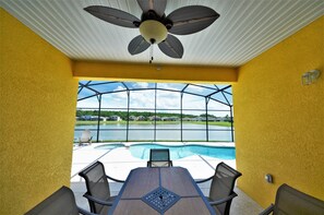 Eine überdachte Terrasse mit Tisch und Stühlen, einem Deckenventilator und Blick auf einen Swimmingpool und Häuser.