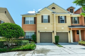A two-story residential building with a beige facade, two garage doors, and a well-maintained front yard with a large tree and trimmed bushes. - Reunion Resort and Spa 835 (Kissimmee)