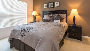 Un dormitorio con cama, dos mesitas de noche con lámparas, una ventana con persianas y tres cuadros enmarcados en la pared.