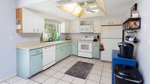 Cocina compacta con armarios blancos y azul claro, microondas, estufa y refrigerador. Hay un ventilador de techo, una ventana que proporciona luz natural y una alfombra pequeña en el suelo.