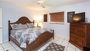 Un dormitorio con cama de madera, ventilador de techo, televisor sobre una cómoda de madera y ventana con persianas.