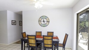 Un comedor con una mesa dispuesta para cuatro personas, rodeada de sillas con respaldo trenzado. Un reloj decorativo está colgado en la pared sobre la mesa.