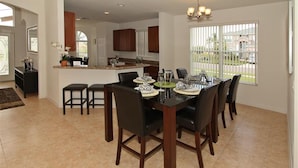 A spacious dining area with a large table and chairs, a kitchen in the background, and a view of the outside through large windows. - Crystal Cove 4725 (Kissimmee)