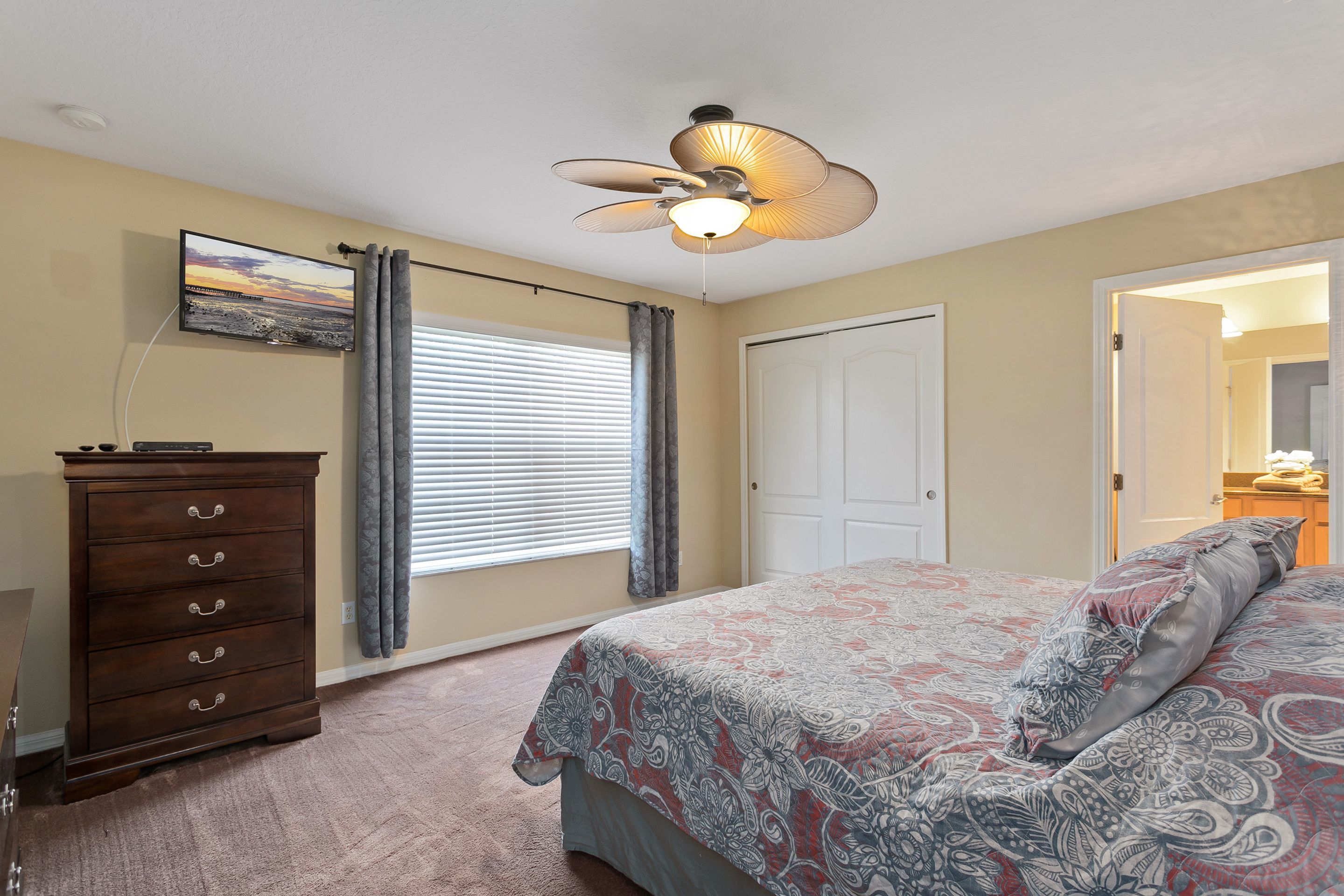 A bedroom with a bed, a dresser, a ceiling fan, a window with curtains, and a door.