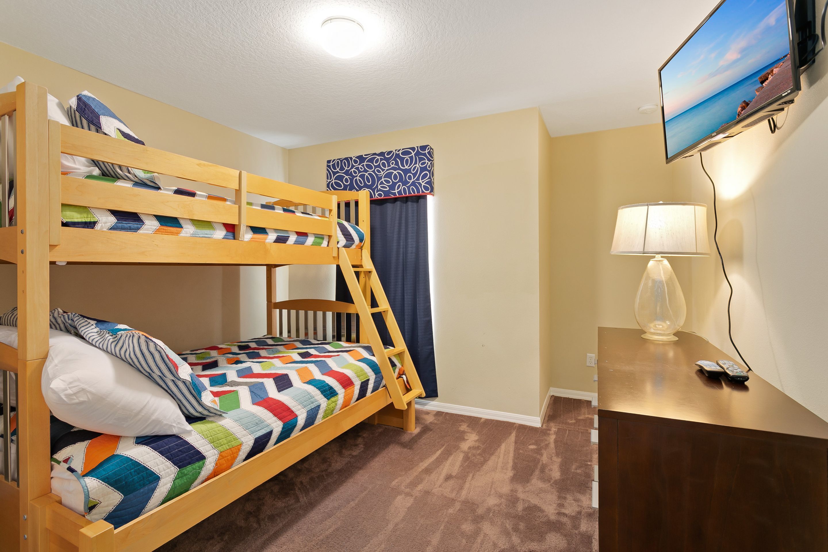 A bunk bed with a colorful quilt, a desk with a lamp, and a TV on the wall.