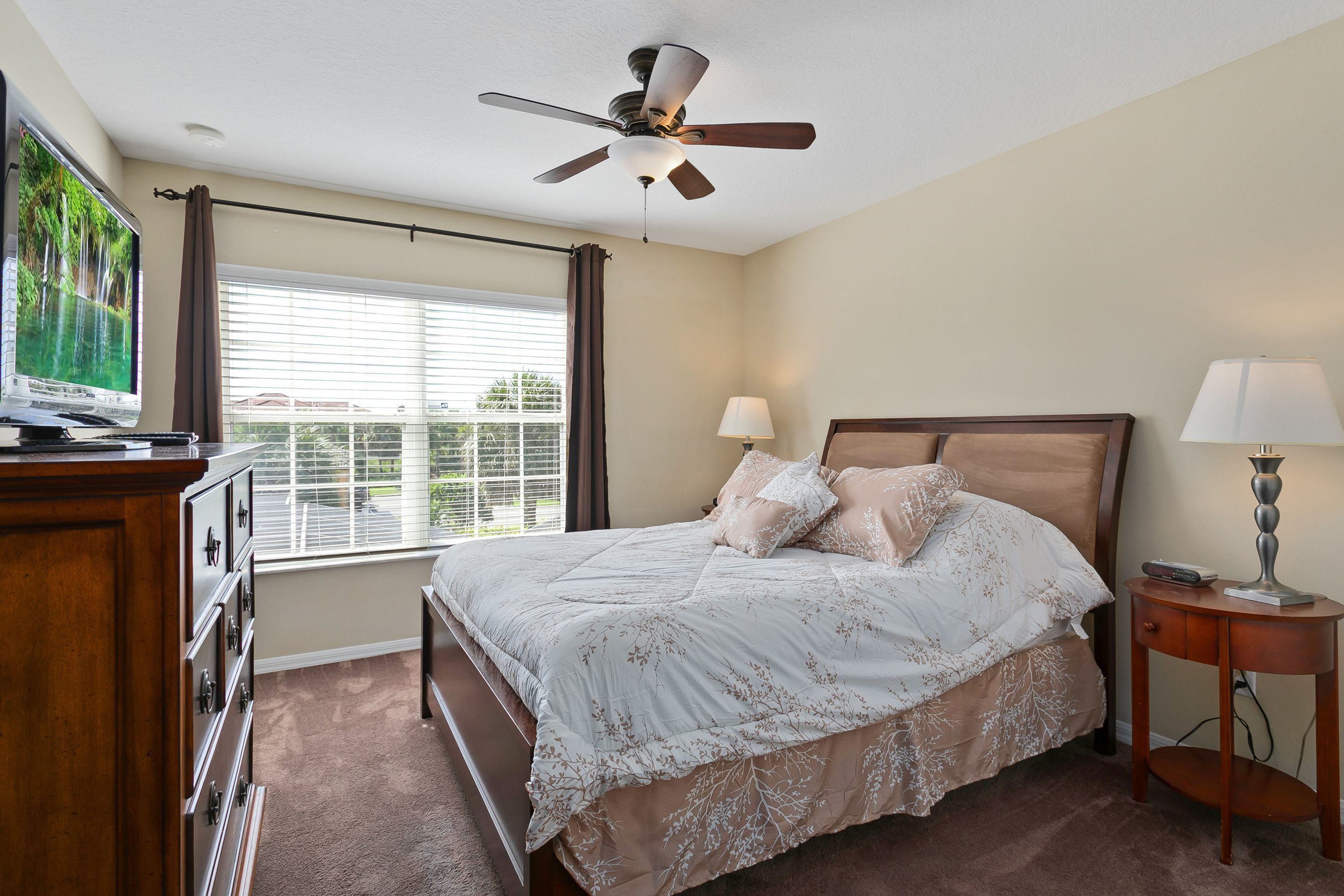 Une chambre à coucher comprenant un lit, un ventilateur de plafond, une télévision, une fenêtre avec des rideaux et une table de chevet avec une lampe.
