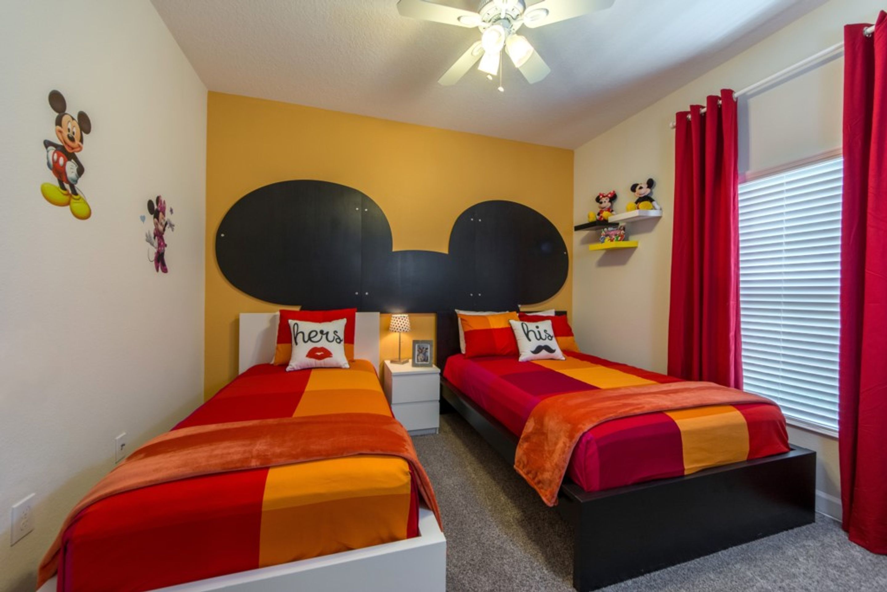 Une chambre avec deux lits, chacun doté d’une tête de lit en forme de tête de Mickey Mouse, et agrémentée de draps et de couvertures aux couleurs rouge et orange, ainsi que d’autocollants muraux à l’effigie de Mickey Mouse.