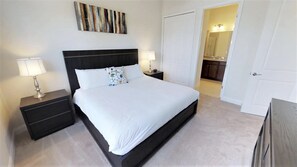 A neatly arranged bedroom with a large bed, two bedside tables with lamps, a decorative wall piece, and a view into the bathroom.