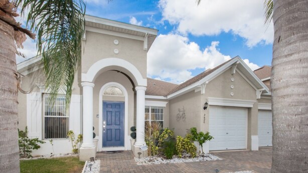 A two-story house with a blue front door, a garage, and a landscaped front yard with palm trees and shrubs. - Sand Hill 2620 (Davenport)