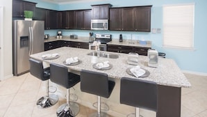 A modern kitchen with dark wood cabinets, a stainless steel refrigerator, a microwave, and a granite countertop. There are four bar stools and a large island with a sink. - Windsor at Westside 2145 (Kissimmee)