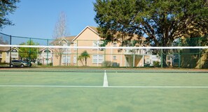 A tennis court with a net, surrounded by a green fence, trees, and a building in the background. - Sweetwater Club 2494 - 55 (Kissimmee)
