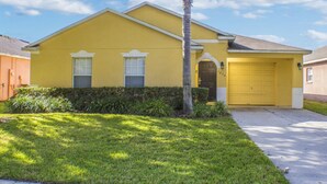 A yellow single-story house with a garage, a front yard with grass and a palm tree, and a clear blue sky. - Silver Creek 1435 (Clermont)