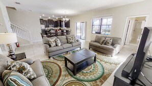 A spacious living room with two sofas, a coffee table, and a TV. There is a kitchen in the background with dark cabinets and a dining area. - Champions Gate 9035 (Davenport)