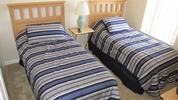 Two single beds with striped bedding, a small bedside table with a lamp, and a window with blinds.