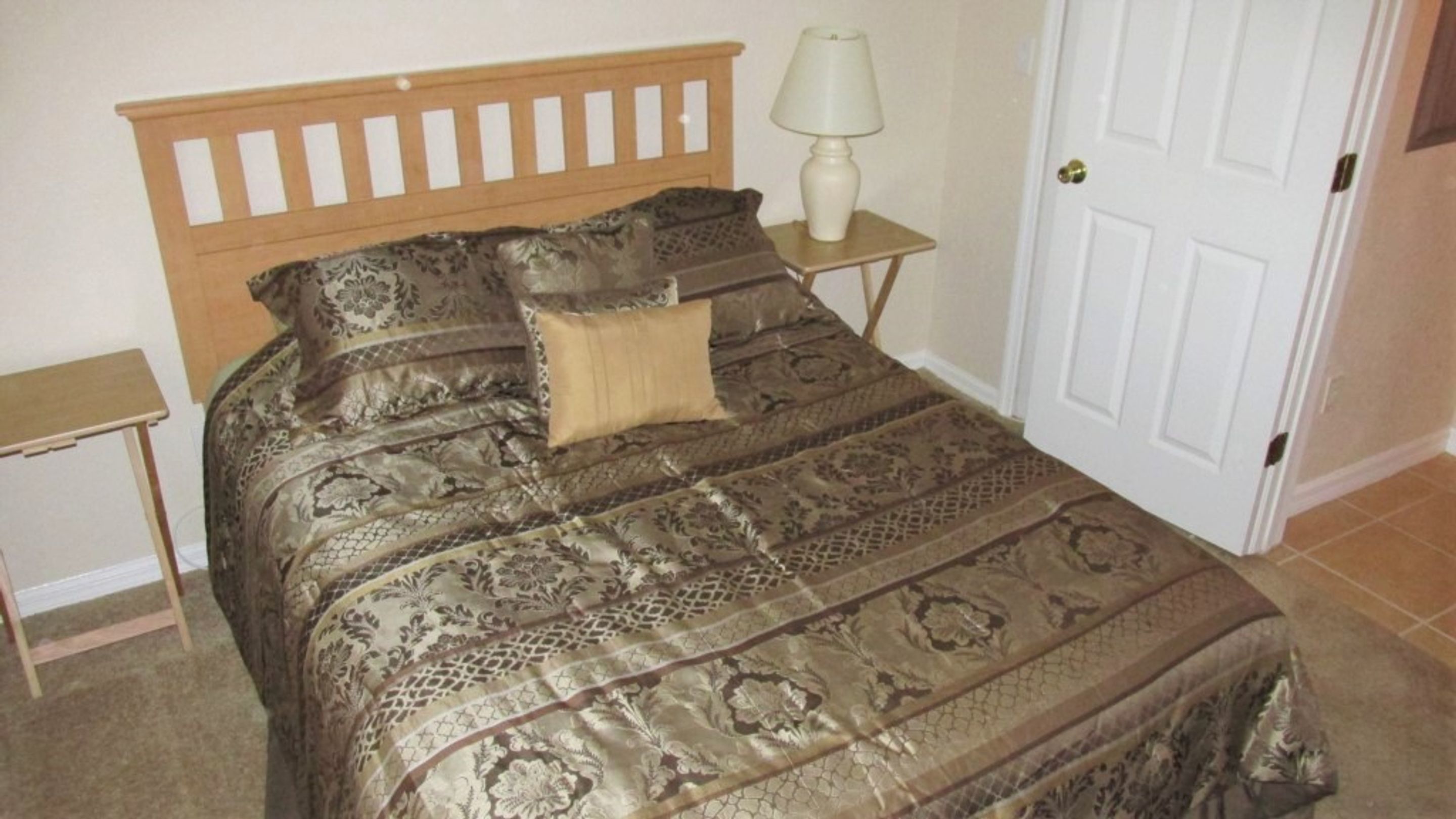 A bedroom with a wooden headboard, a patterned bedspread, a bedside lamp, and a door.