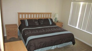 A bedroom with a bed, a nightstand, a lamp, and a window with blinds. - Regal Oaks 2715 (Kissimmee)