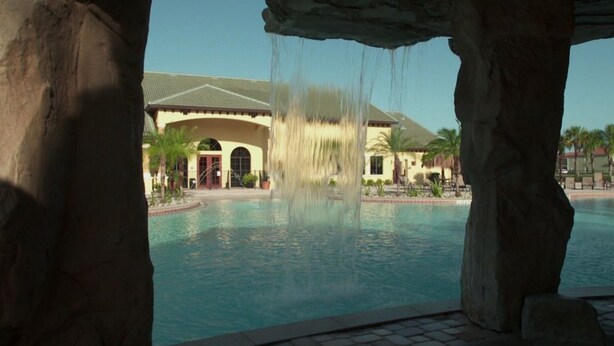 Una piscina con cascada, un edificio con ventanas arqueadas y palmeras al fondo.