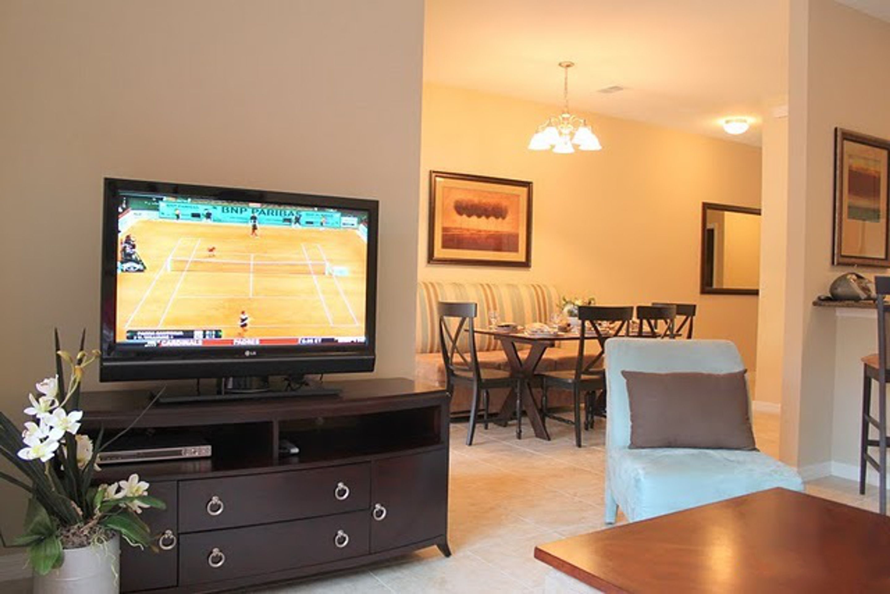 Un salon avec un téléviseur diffusant un match de tennis, un coin repas avec une table et des chaises, et un canapé.