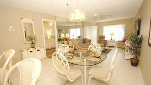 Un coin repas avec une table en verre entourée de chaises blanches, un lustre au-dessus et un ventilateur de plafond.