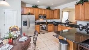 A kitchen with wooden cabinets, stainless steel appliances, a dining table with chairs, and a trash can. - Windsor Hills 2548 (Kissimmee)
