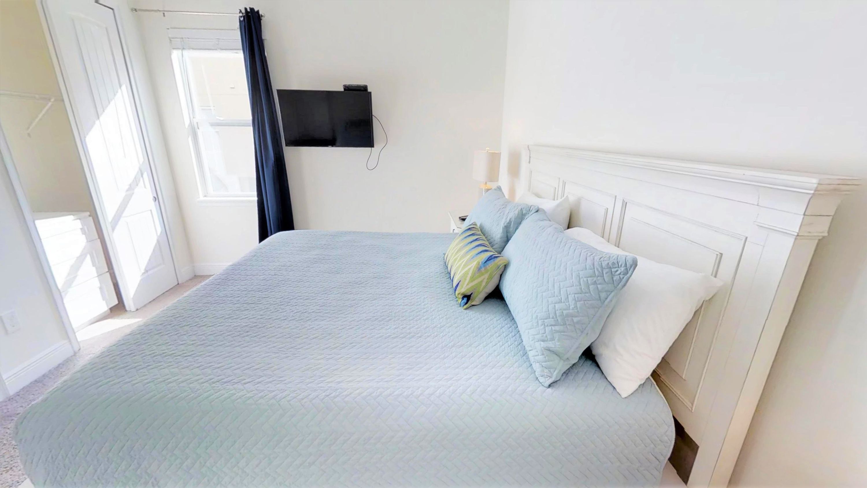 A bedroom with a bed, a TV mounted on the wall, and a window with curtains.
