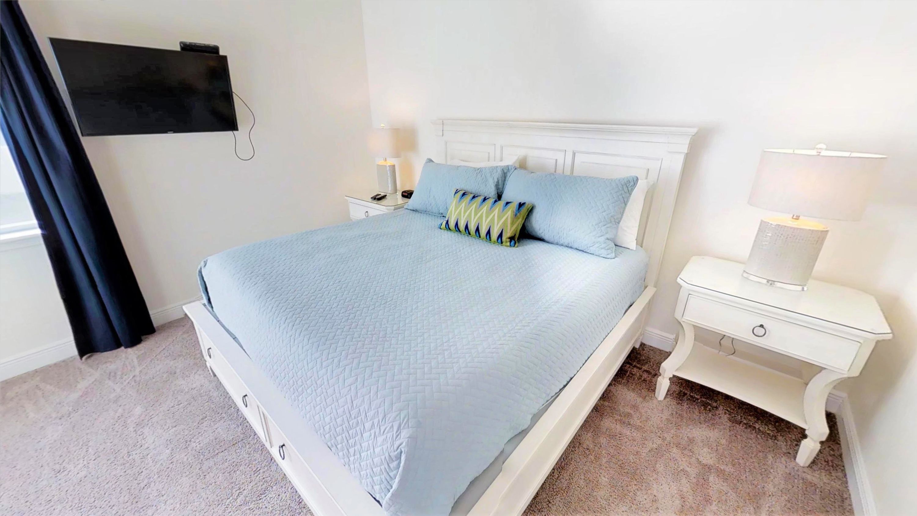 A neatly made bed with a quilted blue bedspread and two pillows, a white nightstand with a lamp, and a wall-mounted TV.