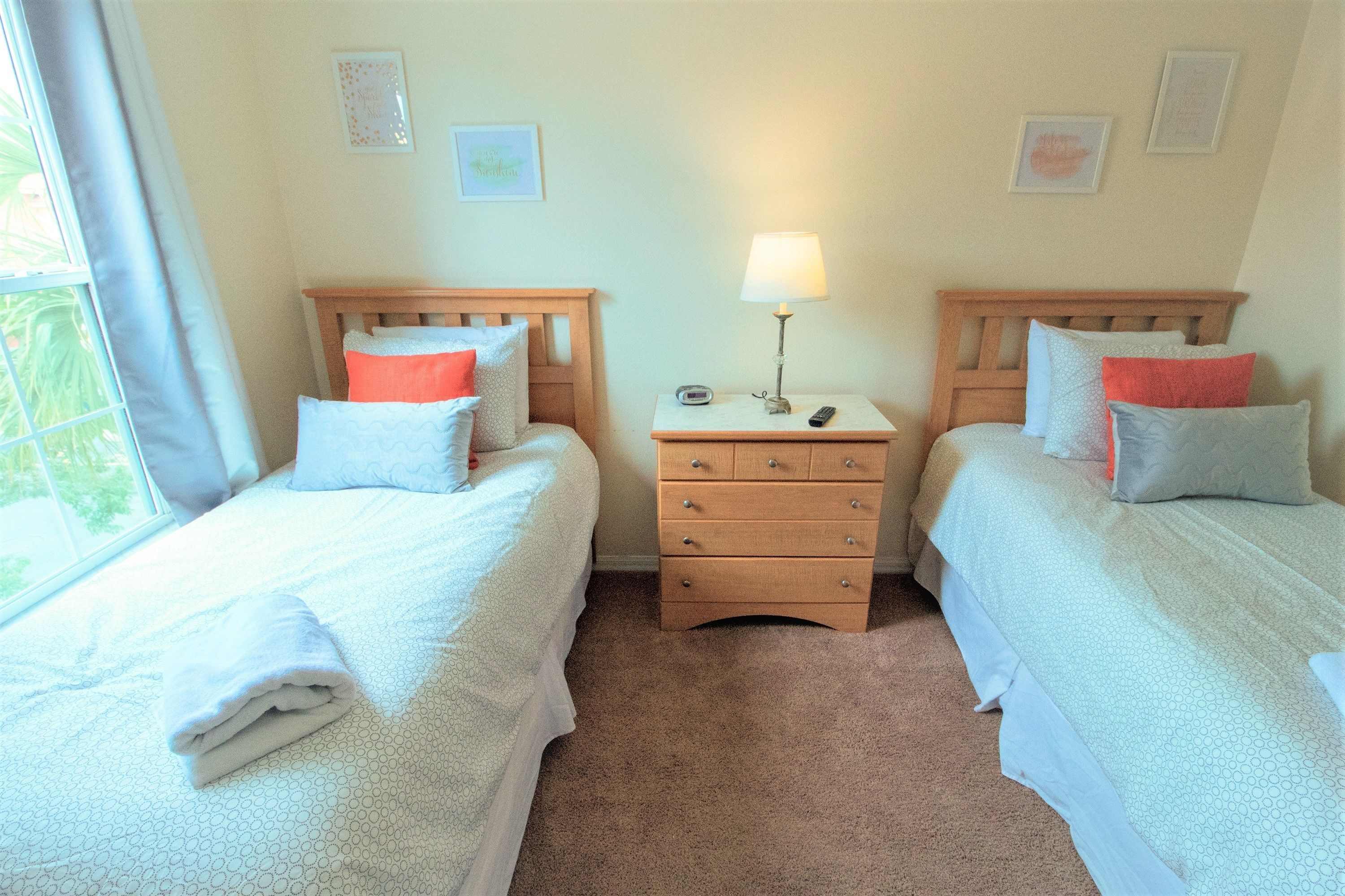 A room with two beds, each with a wooden headboard, a bedside table with a lamp, and a small drawer unit.
