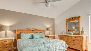 Un dormitorio con cabecera de madera, cama con ropa de cama estampada, dos mesitas de noche con lámparas, cómoda con cajones y un espejo grande.