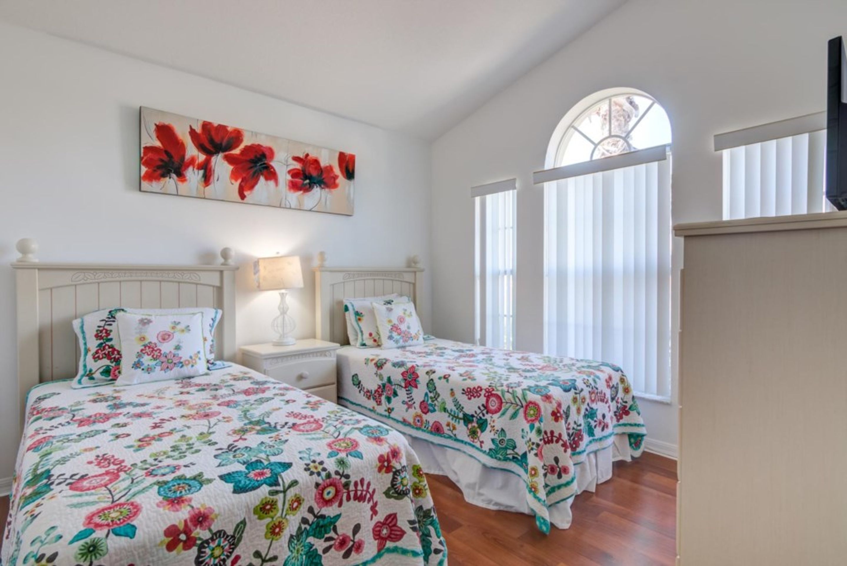 A bedroom with two beds, a nightstand, a lamp, a floral painting, and a window with blinds.