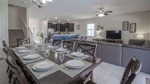 A dining area with a long table set for six, a kitchen in the background, and a living area with a sofa and TV. - Champions Gate 1476 (Davenport)