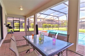 A covered patio with a glass table, chairs, and a view of a swimming pool.