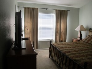 A bedroom with a bed, a TV on a stand, a window with curtains, and a nightstand with a lamp. - Windsor Hills 2503 (Kissimmee)