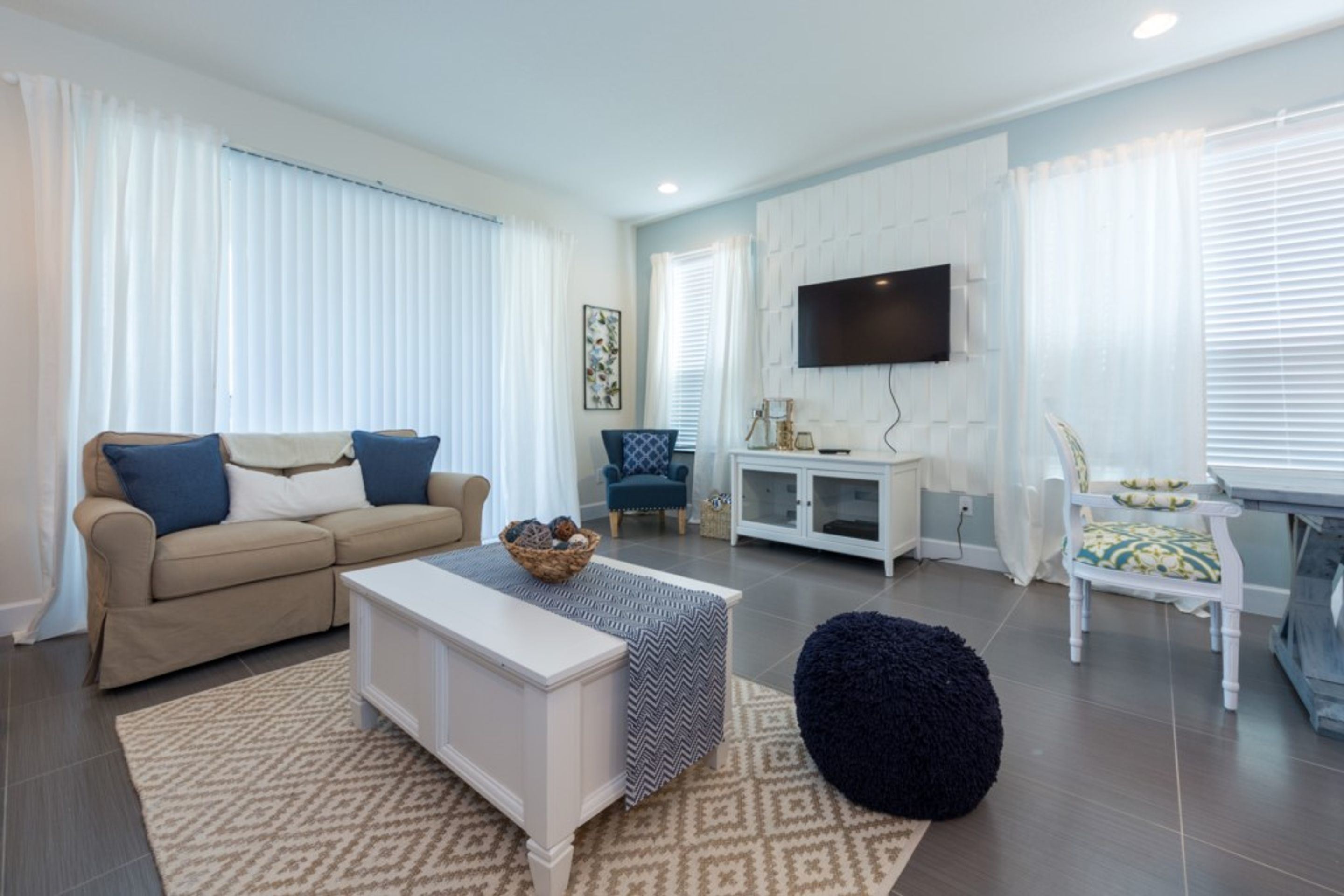 A modern living room with a beige sofa, a white coffee table, and a black pouf. There is a flat-screen TV mounted on the wall and a white cabinet with a glass top.