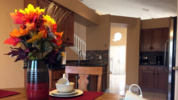 A modern kitchen with wooden cabinets, a dining table with red placemats, and a vase of colorful flowers. - Chatham Park 2718 (Kissimmee)