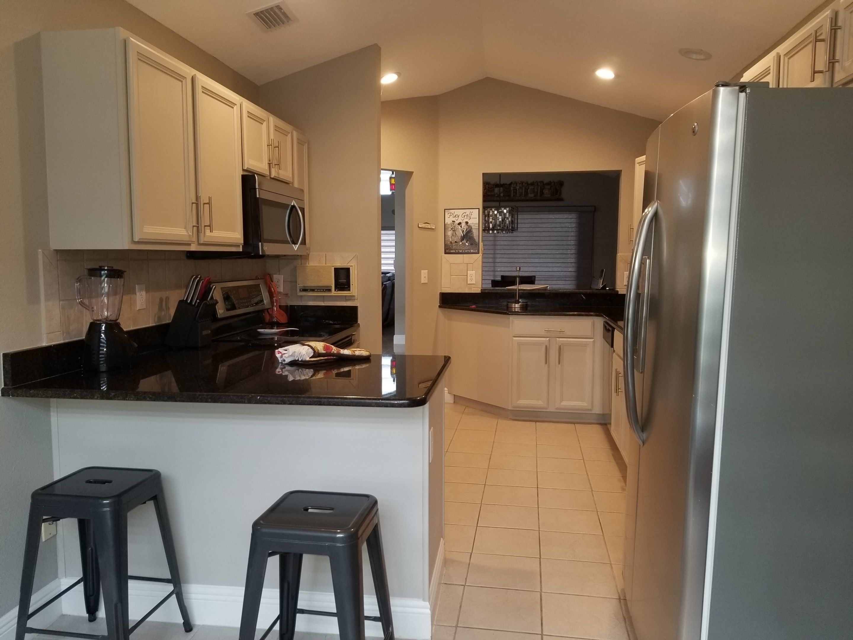 A kitchen with a refrigerator, microwave, and a blender on the counter. There are two stools and a small island.